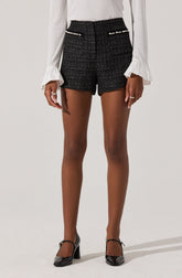 A woman is wearing high-waisted black tweed shorts with a subtle pattern, featuring decorative pearl embellishments on the front pockets. She is also dressed in a long-sleeved white blouse with ruffled cuffs. She completes the outfit with black patent leather high heels with ankle straps.